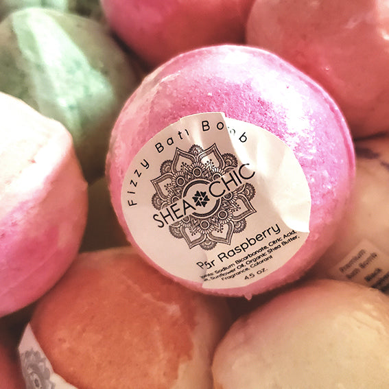 A bath bomb by Shea Chic with a pink, glittery exterior. The label on the bomb reads 'Organic Shea Butter Bath Bomb' with the brand name 'Shea Chic' and the scent 'Raspberry'.
